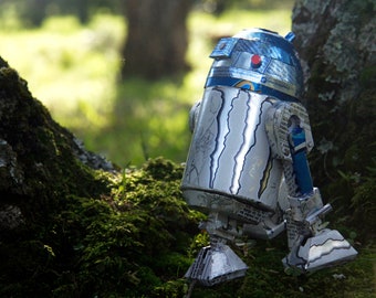 May include: A silver and blue robot made of recycled materials, standing in a forest setting. The robot is facing away from the camera and has a round head with a red button.