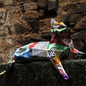 May include: A colorful cat sculpture made from recycled materials, including food packaging. The cat is lying down with its head raised and its tail curled around its body. The sculpture is made from a variety of materials, including aluminum, plastic, and cardboard. The cat is a vibrant mix of colors, including red, green, blue, and yellow.
