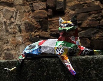 May include: A colourful cat sculpture made from recycled materials, including food packaging. The cat is lying down with its head raised and its tail curled around its body. The sculpture is made from a variety of materials, including aluminium, plastic, and cardboard. The cat is a vibrant mix of colours, including red, green, blue, and yellow.