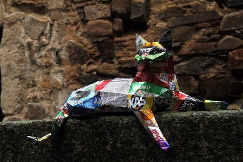 May include: A colorful cat sculpture made from recycled materials, including food packaging. The cat is lying down with its head raised and its tail curled around its body. The sculpture is made from a variety of materials, including aluminum, plastic, and cardboard. The cat is a vibrant mix of colors, including red, green, blue, and yellow.