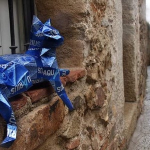 May include: A blue cat sculpture made from recycled materials, sitting on a brick wall. The sculpture is made from blue plastic and has the words "Aquarius" and "Smaqu" printed on it.