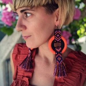 May include: A woman with short blonde hair wearing a red top and large orange and black tassel earrings. The earrings have a geometric pattern and a large orange circle.