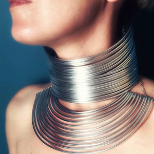 Könnte beinhalten: Eine silberne Choker-Kette mit mehreren Lagen dünner Metallringe. Die Kette wird von einer Person mit nach unten geneigtem Kopf getragen.