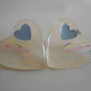 Op de afbeelding: Een paar transparante hartvormige oorbellen met een blauw hart en gele, roze en blauwe lijnen in elk hart.