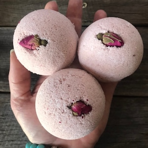 May include: Three pink bath bombs with a textured surface, each with a dried rose bud nestled in a small indentation. The bath bombs are held in a hand, with a wooden background.