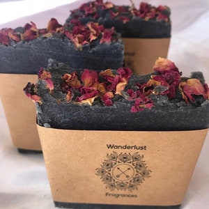 Puede incluir: Tres jabones de color carbón vegetal cubiertos con pétalos de rosa secos. Cada pastilla está envuelta en una funda de papel marrón con las palabras "Wanderlust Fragrances" y un logotipo decorativo. Aspecto rústico y natural.