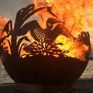 May include: A metal fire pit shaped like a sphere with a cut-out design of a loon and reeds. The fire pit is burning with orange flames and the text "FIREBALL" is visible on the base.