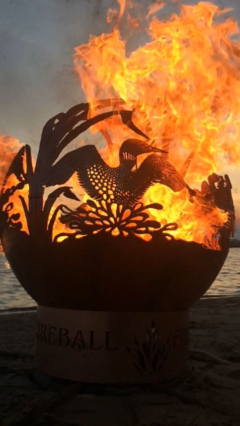 May include: A metal fire pit shaped like a sphere with a cut-out design of a loon and reeds. The fire pit is burning with orange flames and the text "FIREBALL" is visible on the base.