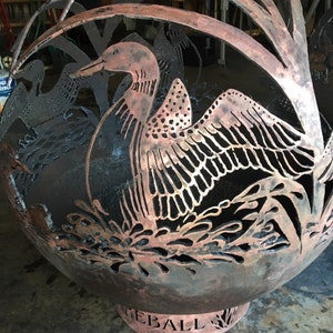 May include: A large, round, metal fire pit with a cut-out design of a loon bird. The fire pit is copper colored and has the word "FIREBALL" inscribed on the base.