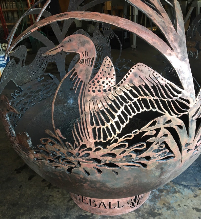 May include: A large, round, metal fire pit with a cut-out design of a loon bird. The fire pit is copper colored and has the word "FIREBALL" inscribed on the base.