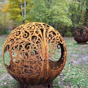 May include: A large, rusty metal sphere with a decorative, intricate design cut into the surface. The sphere is sitting on a grassy surface with fallen leaves.