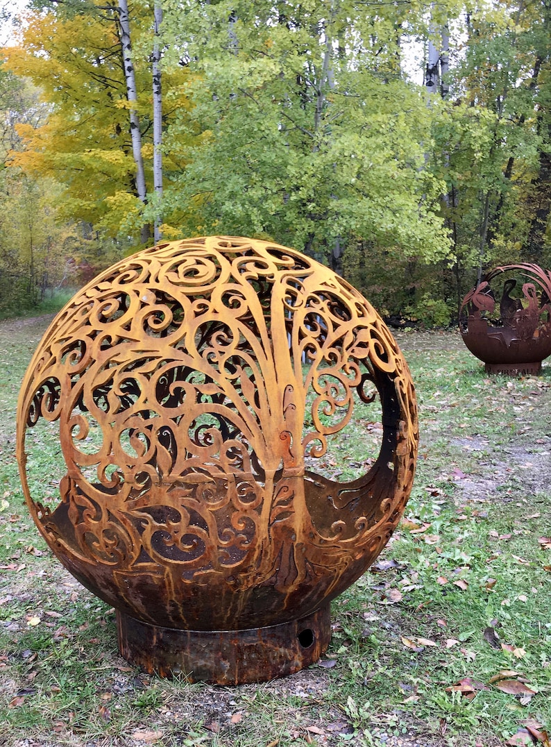 May include: A large, rusty metal sphere with a decorative, intricate design cut into the surface. The sphere is sitting on a grassy surface with fallen leaves.