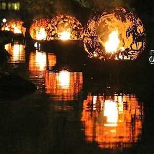 May include: A series of metal fire orbs with intricate designs are floating on a dark body of water. The orbs are lit from within, casting a warm glow on the water's surface.