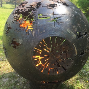 May include: A large, round, metal fire pit with a decorative design. The fire pit is made of steel and has a black finish. The fire pit has a large opening in the center, which is surrounded by a ring of smaller openings. The fire pit is sitting on a grassy surface.