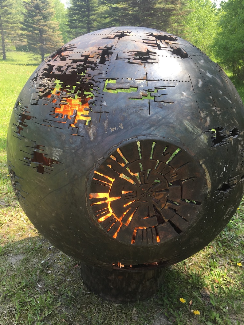 May include: A large, round, metal fire pit with a decorative design. The fire pit is made of steel and has a black finish. The fire pit has a large opening in the center, which is surrounded by a ring of smaller openings. The fire pit is sitting on a grassy surface.