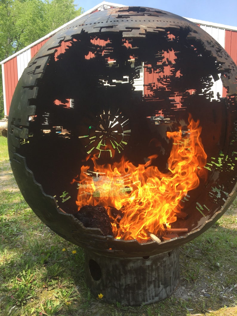 May include: A large metal sphere with a cut-out design resembling the Death Star from Star Wars. The sphere is sitting on a metal base and has a fire burning inside.