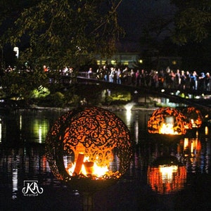 May include: Two large, ornate, metal spheres with flames burning inside, floating on a dark body of water at night. The spheres are decorated with intricate designs and are illuminated by the flames.