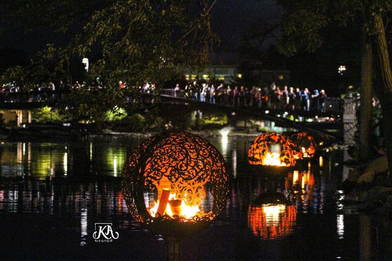 May include: Two large, ornate, metal spheres with flames burning inside, floating on a dark body of water at night. The spheres are decorated with intricate designs and are illuminated by the flames.