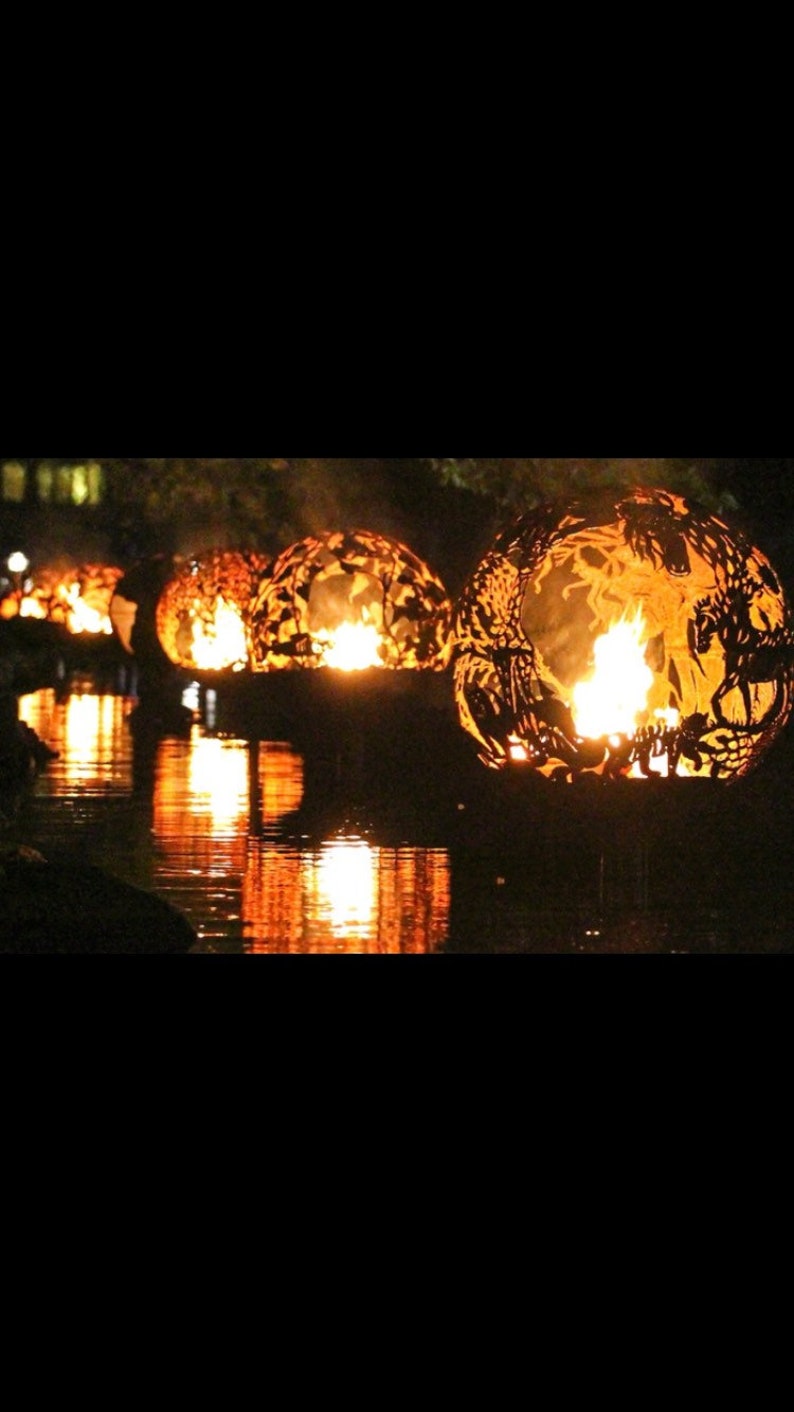 May include: Several large, round, metal fire orbs with intricate cut-out designs are burning brightly in a dark body of water. The orbs are floating on the water and casting a warm glow on the surrounding area.