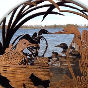 May include: A rusty metal sculpture of a family of loons with their young, set against a backdrop of water and reeds. The sculpture is in the shape of a sphere.
