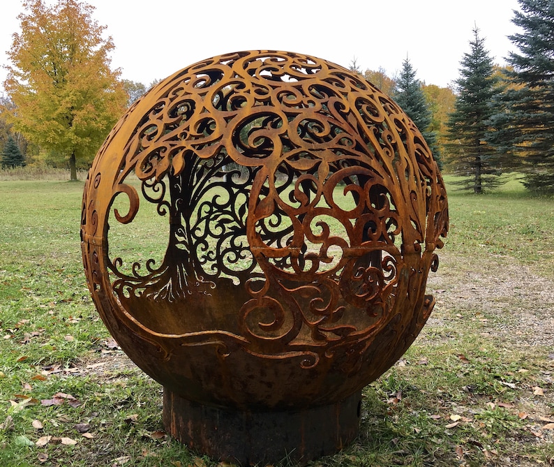 May include: A large, rusty metal sphere with a cut-out design of a tree with intricate branches and leaves. The sphere is sitting on a grassy surface.