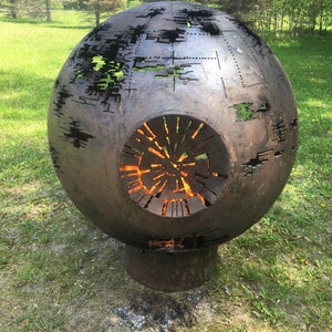 May include: A large, round, metal fire pit shaped like the Death Star from Star Wars. The fire pit is made of metal and has a large opening in the front. The fire pit is sitting on a grassy lawn.