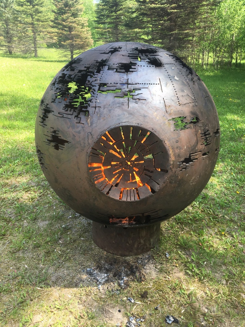 May include: A large, round, metal fire pit shaped like the Death Star from Star Wars. The fire pit is made of metal and has a large opening in the front. The fire pit is sitting on a grassy lawn.