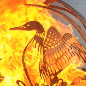 May include: A metal fire pit shaped like a loon with its wings spread. The fire pit is lit and the flames are visible through the cutouts in the metal. The loon is sitting on a bed of water lilies.