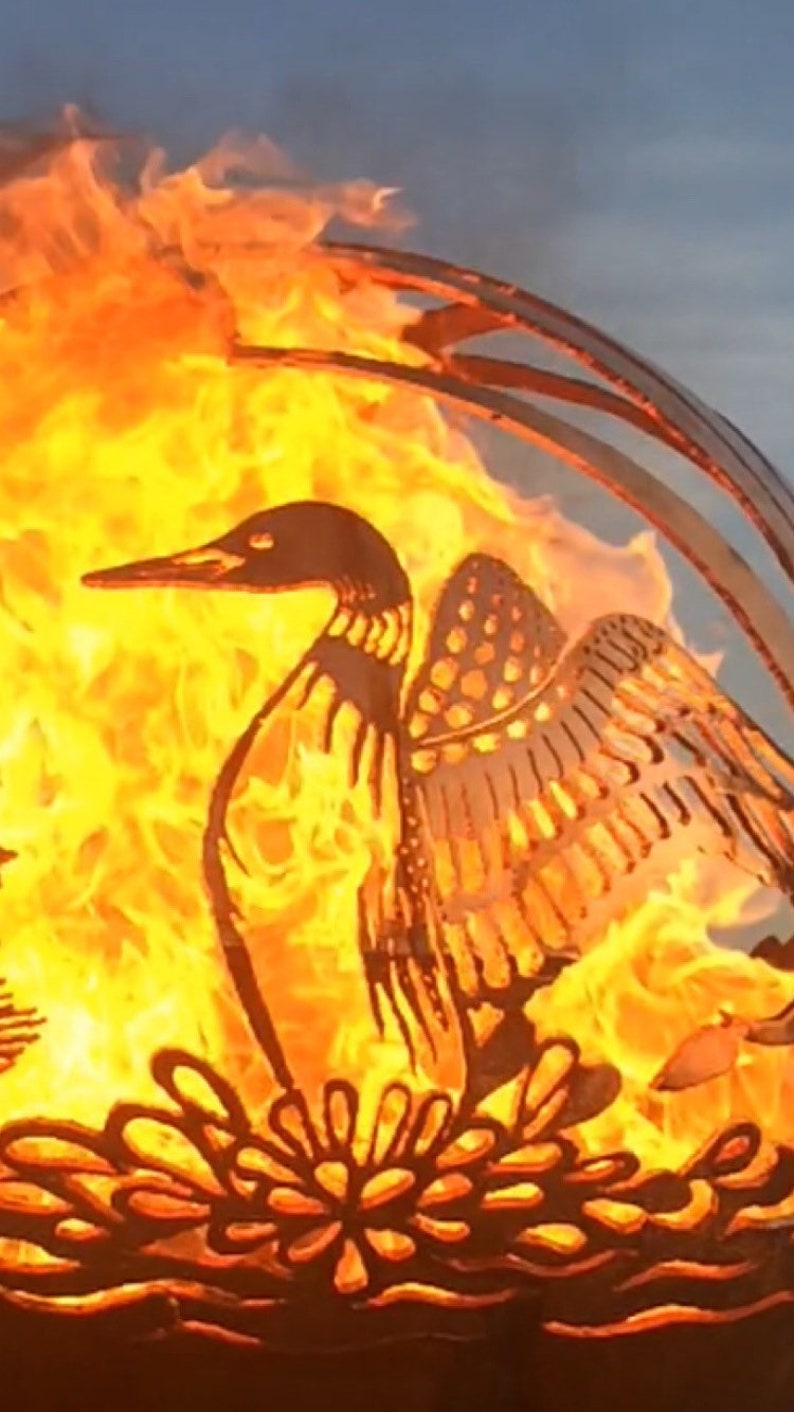 May include: A metal fire pit shaped like a loon with its wings spread. The fire pit is lit and the flames are visible through the cutouts in the metal. The loon is sitting on a bed of water lilies.