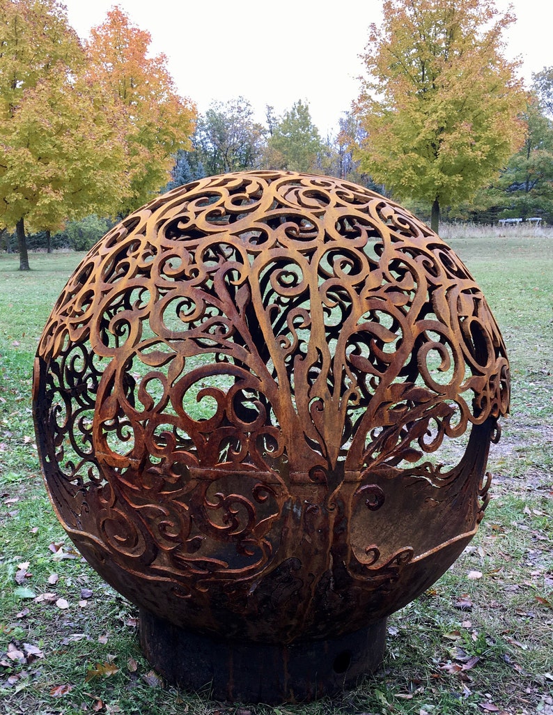 May include: A large, rusty metal sphere with a decorative, intricate design of swirling branches and leaves. The sphere is sitting on a grassy surface with fallen leaves.