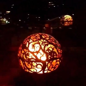 May include: A decorative metal fire pit with a swirling, tree-like design. The fire pit is lit and glowing orange. There are several other fire pits in the background.