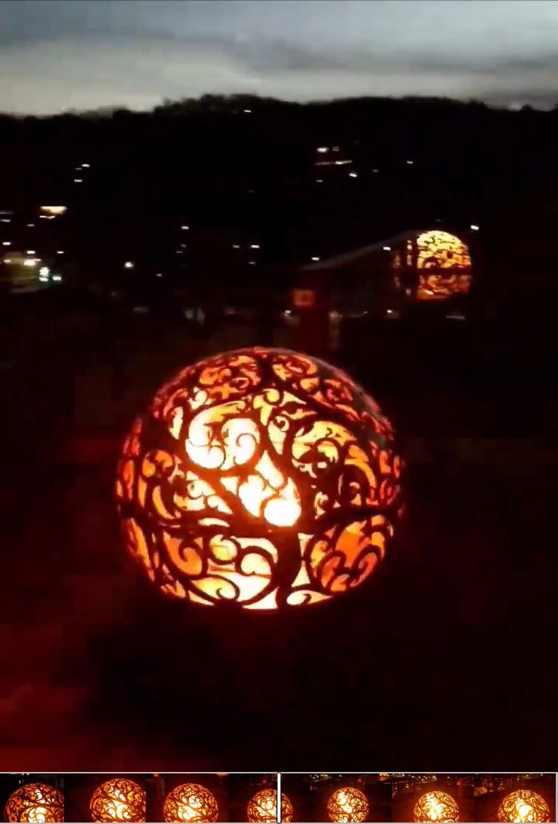 May include: A decorative metal fire pit with a swirling, tree-like design. The fire pit is lit and glowing orange. There are several other fire pits in the background.