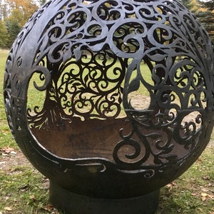 May include: A large, black metal sphere with a decorative, cut-out design resembling a tree. The sphere is sitting on a grassy surface with fallen leaves.