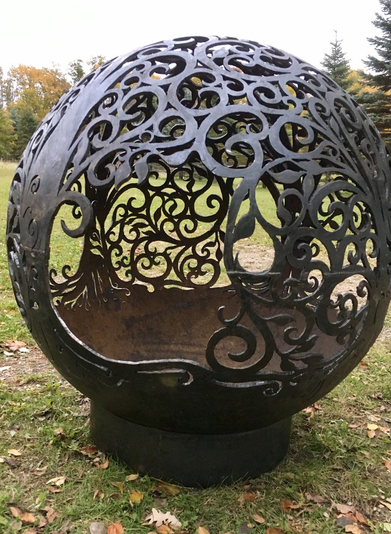 May include: A large, black metal sphere with a decorative, cut-out design resembling a tree. The sphere is sitting on a grassy surface with fallen leaves.