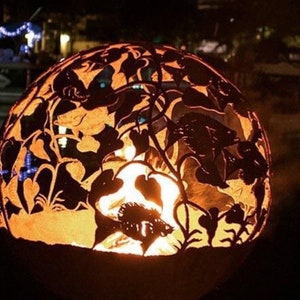 May include: A metal fire pit with a round, open design. The fire pit is decorated with a cut-out pattern of flowers and leaves. The fire pit is lit and glowing orange.