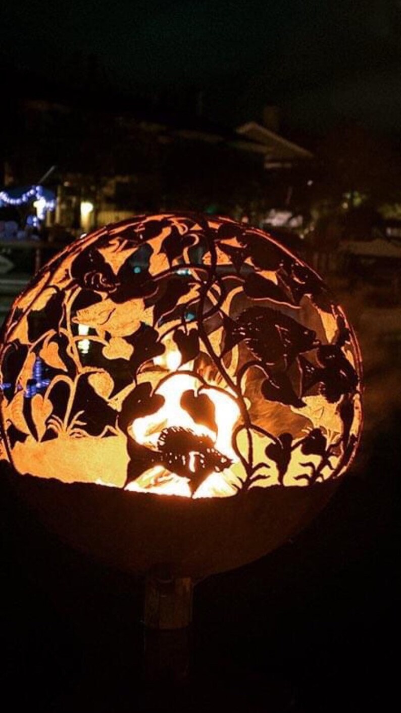 May include: A metal fire pit with a round, open design. The fire pit is decorated with a cut-out pattern of flowers and leaves. The fire pit is lit and glowing orange.