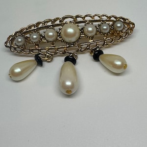 May include: A vintage gold-tone brooch with faux pearls of different sizes. The brooch has an openwork frame and three dangling pear-shaped pearl drops with black accents. A classic accessory for a formal occasion.