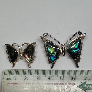 Puede incluir: Dos broches de mariposa de plata con alas iridiscentes. El broche más grande mide aproximadamente 4 cm de ancho y el más pequeño aproximadamente 2 cm de ancho.