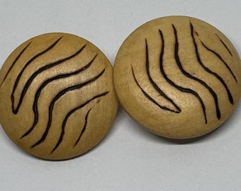 May include: A pair of round, light brown earrings with a wood grain effect. Each earring has a series of dark brown, curved lines across its surface. The earrings are set against a plain, light background.