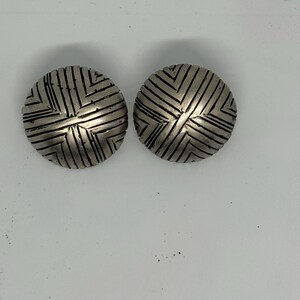 May include: Two silver-toned, round stud earrings. Each earring features a textured surface with a geometric, linear pattern. The earrings are displayed on a white surface, showcasing their design and metallic finish.