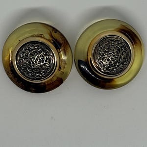 May include: A pair of large, round clip-on earrings. The earrings have a gold-colored metal rim and a central, ornate silver-colored design. The outer portion of the earrings is a translucent amber color with brown marbling.