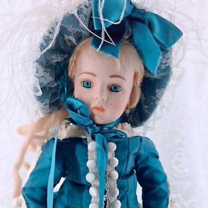 May include: A porcelain doll with blonde hair and blue eyes wearing a teal satin jacket with white buttons, a matching teal bonnet with lace trim and a large bow, and cream-colored lace accents on the sleeves and jacket edges.
