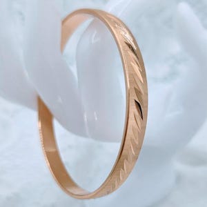 Op de afbeelding: Een roségouden armband met een gepolijste afwerking en een herhalend diagonaal geëtst patroon op het buitenoppervlak. De metalen band heeft een slank, cirkelvormig profiel met een constante breedte over de gehele omtrek.