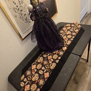 以下が含まれることがあります： 緑色の肌をした装飾用の魔女の人形で、濃い紫と黒のレースのドレスを着用し、杖の上に銀色のキラキラした球体を持っています。人形は、さまざまなハロウィーンをテーマにした円形のラベルが繰り返される模様の黒いテーブルランナーの上に立っています。