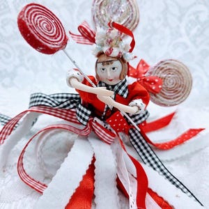 Puede incluir: Una caprichosa figura de muñeca vestida con un atuendo rojo y negro, sosteniendo un caramelo en espiral rojo y blanco. La muñeca tiene un sombrero decorativo y está rodeada de cintas rojas, blancas y negras y caramelos adicionales, creando una apariencia festiva y artesanal.