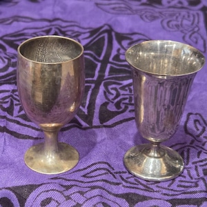 May include: Two silver-toned metal goblets with stems and circular bases. The left goblet has a smooth, rounded bowl, while the right goblet features a slightly flared rim and vertical fluted detailing along the exterior of the cup.