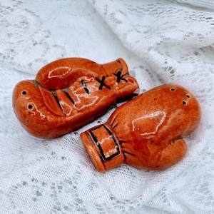 Puede incluir: Un par de saleros y pimenteros con forma de guantes de boxeo de cerámica. Los guantes son de color marrón anaranjado brillante con detalles negros, incluyendo los cordones y las correas de la muñeca. Cada salero tiene pequeños agujeros en la parte superior.