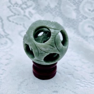 May include: A carved, pale green sphere with intricate floral and geometric cutouts. The sphere sits atop a dark red, round wooden stand. The carving has a smooth, polished finish, and the overall aesthetic is decorative.