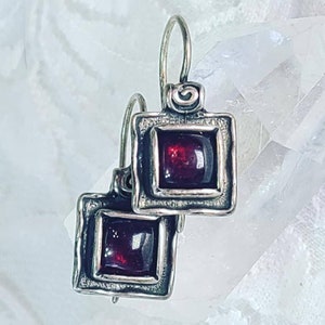 Pode incluir: Um par de brincos quadrados com pedras translúcidas de cor vermelho-escuro engastadas em armações de prata oxidada. Cada brinco apresenta uma borda de metal texturizada e rústica e um gancho curvo para fixação na orelha.