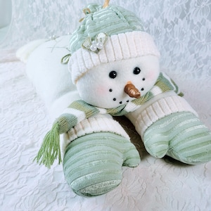 Puede incluir: Decoración de muñeco de nieve de peluche con cuerpo blanco y gorro, bufanda y mitones verdes. El muñeco de nieve tiene una nariz de zanahoria, ojos negros y una sonrisa cosida. El sombrero está adornado con una pequeña flor y perlas.